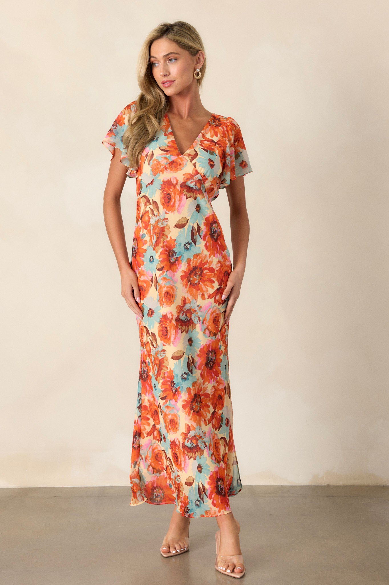 Side view of an orange floral dress showcasing the v-neckline, floral print design, leg slit on the left leg, and the sheer short sleeves.