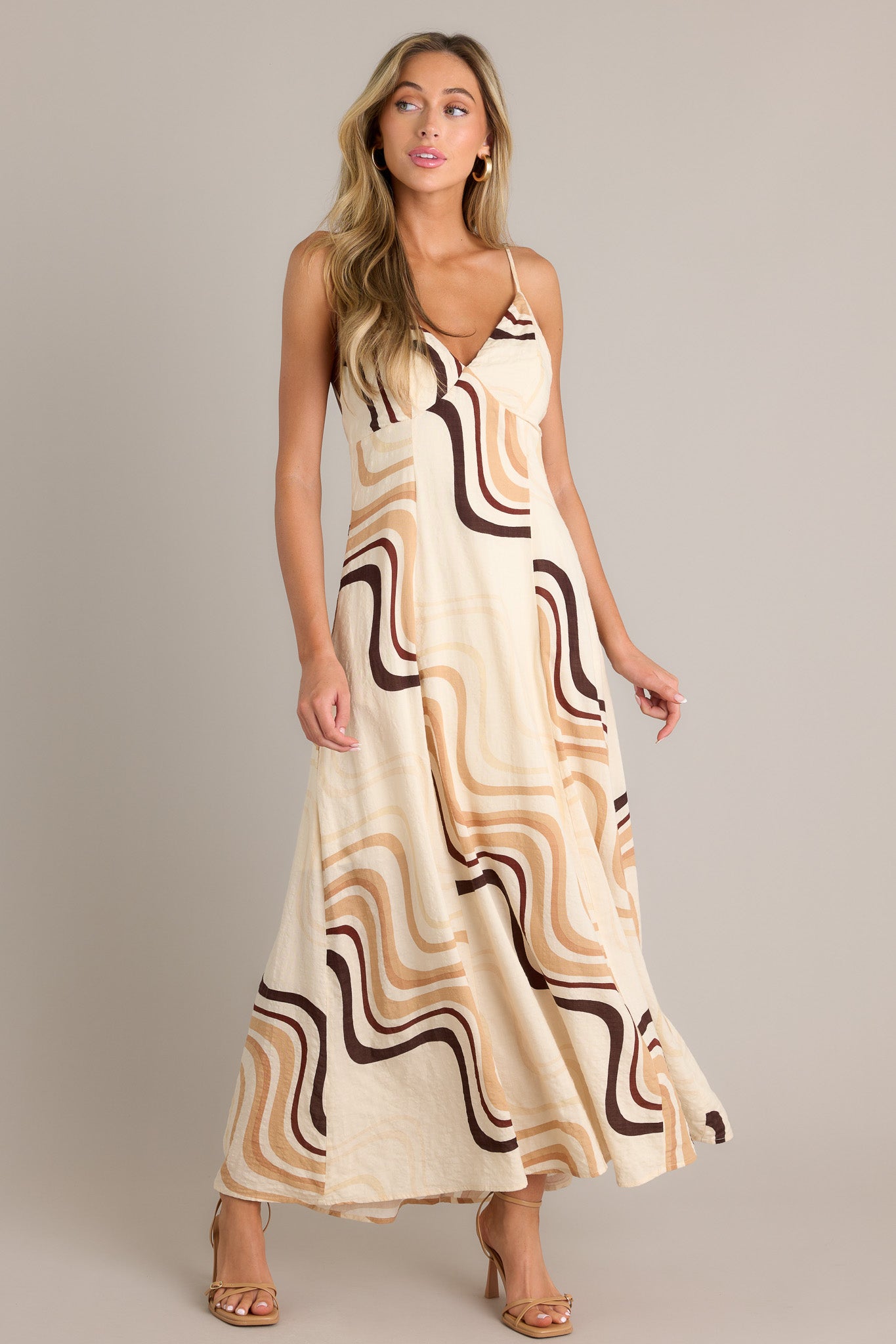 Front angled view of a beige maxi dress featuring a plunging v-neckline, a tie back, buttons down the back, functional hip pockets, and a fun and unique print