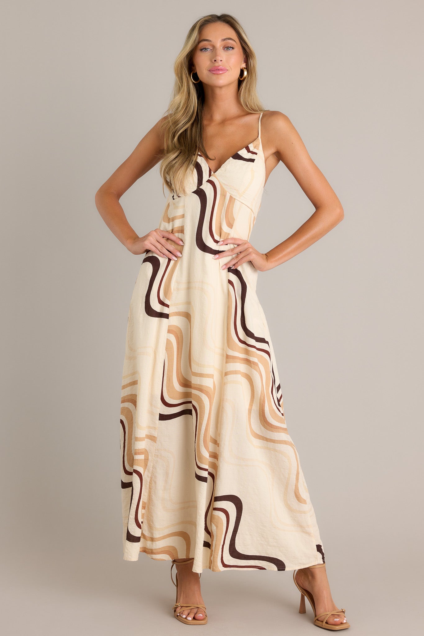 Full length view of a beige maxi dress with a plunging v-neckline, a tie back, buttons down the back, functional hip pockets, and a fun and unique print