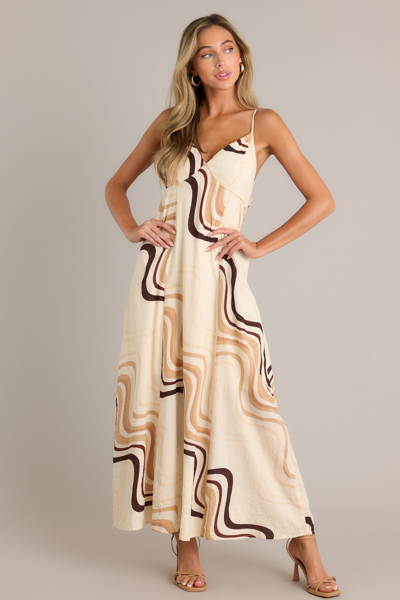 Front view of a beige maxi dress featuring a plunging v-neckline, a tie back, functional hip pockets, and a fun and unique print.