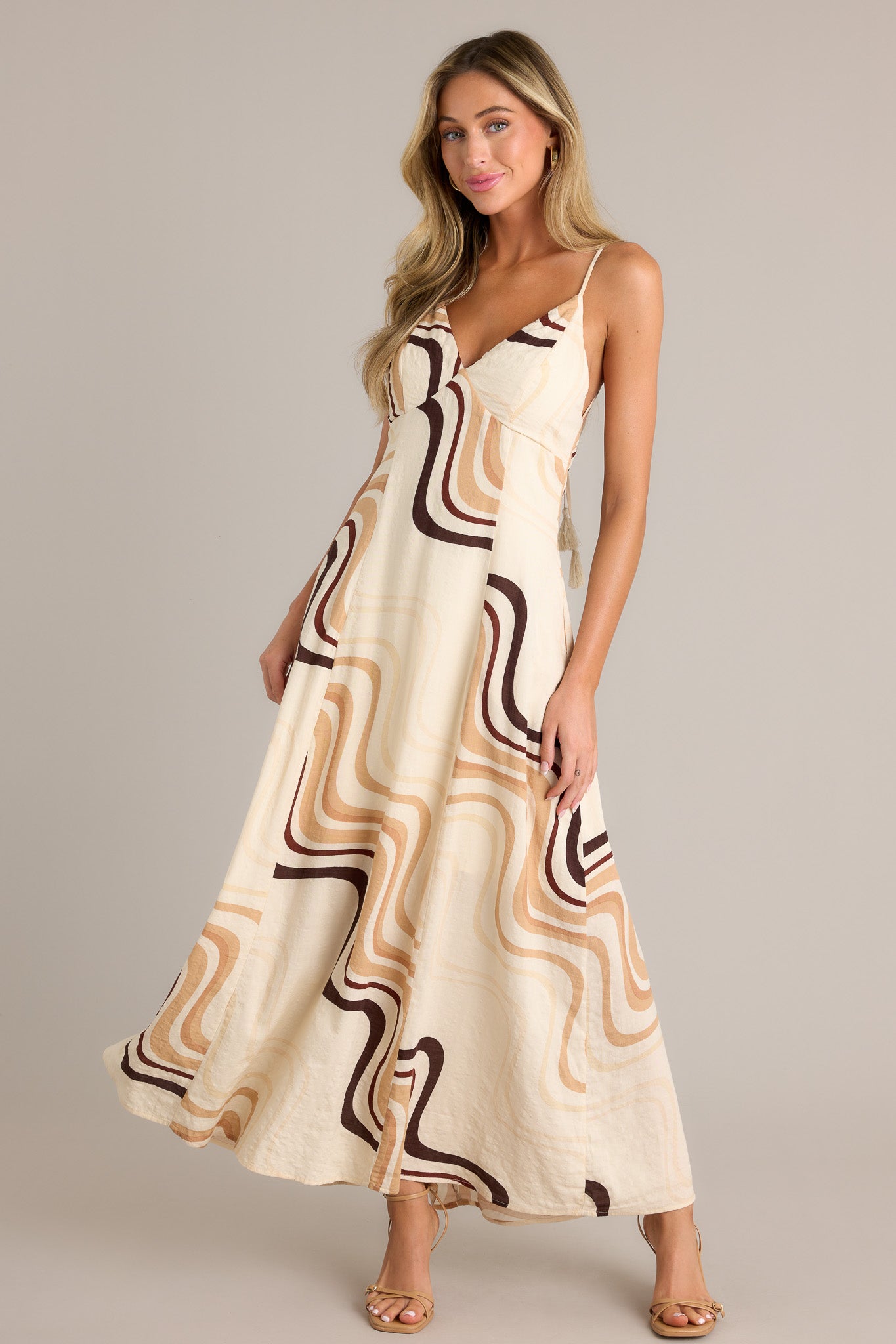 Action shot of a beige maxi dress displaying the fit and movement, highlighting the plunging v-neckline, tie back, buttons down the back, functional hip pockets, and fun and unique print.