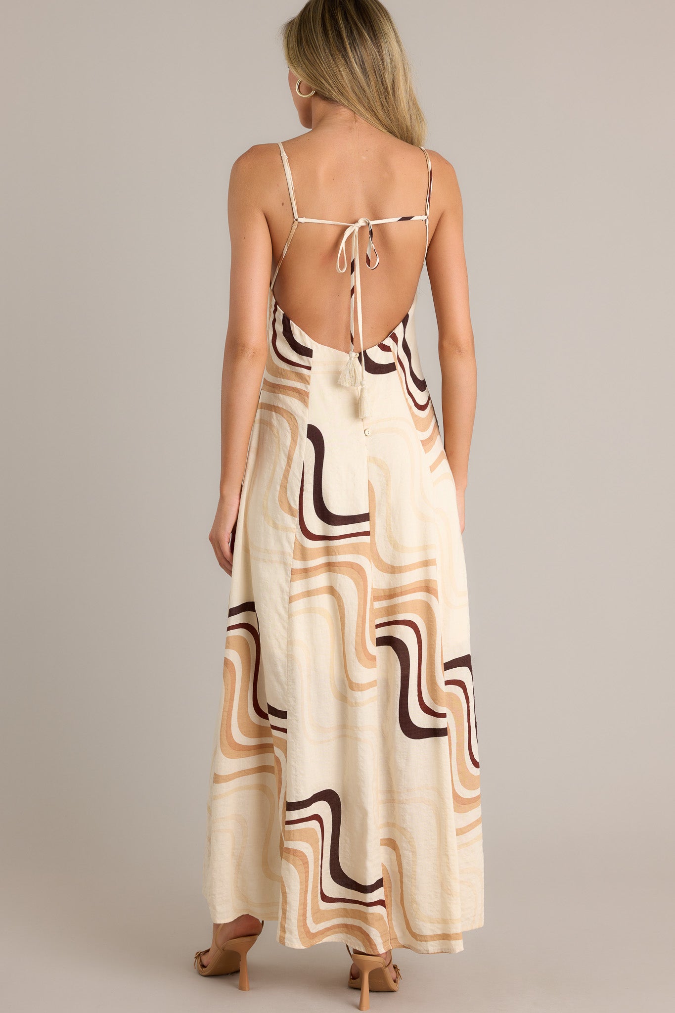 Back view of a beige maxi dress highlighting the tie back, buttons down the back, functional hip pockets, and overall fit.