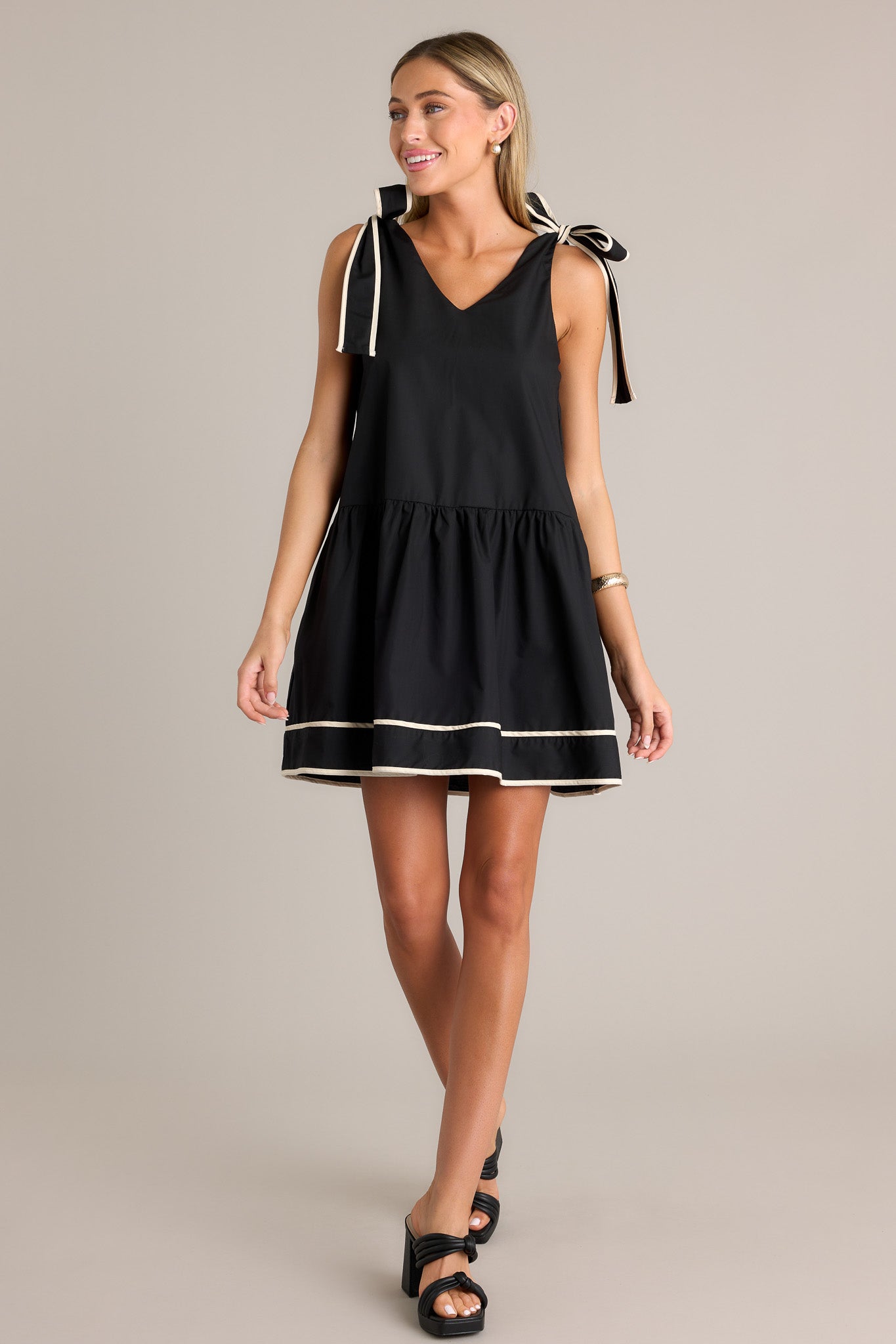 Front view of the black mini dress featuring a single tier and high contrast trim, with self-tie shoulder straps and functional hip pockets.