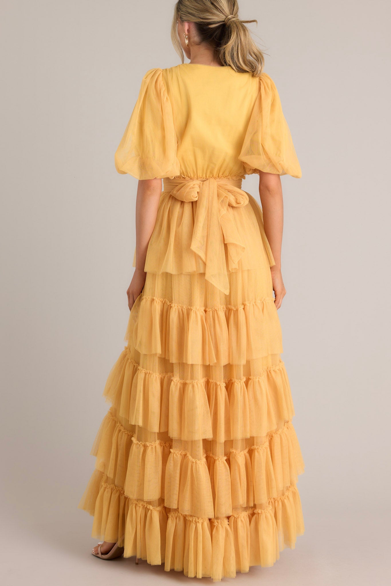 Back view of this yellow dress features a v-neckline, puff sleeves, elastic stretch around the waist, a removable self-tie at the waist, and a tiered flowy skirt.