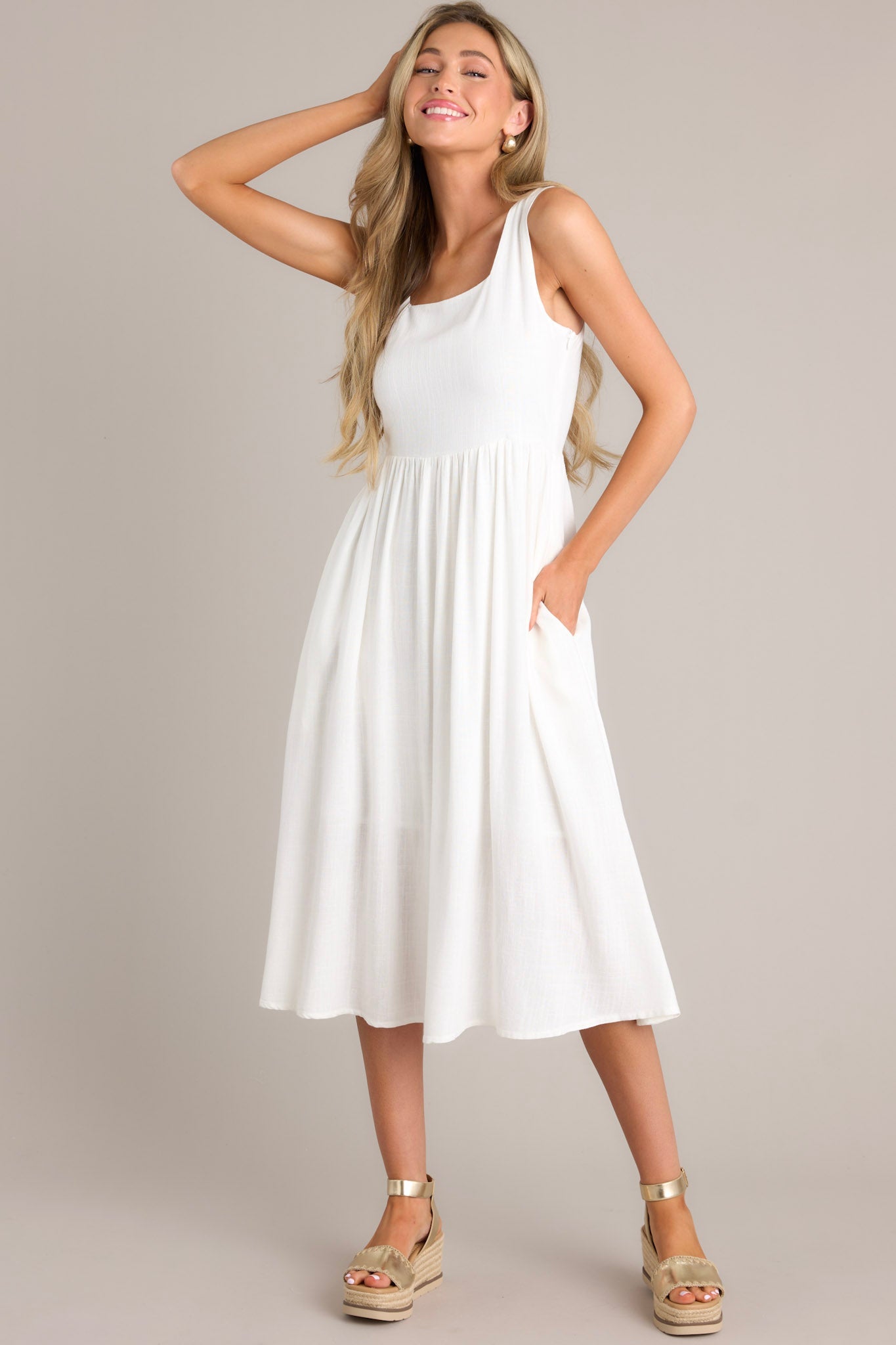 Angled full body view of this white dress with a square neckline, thick straps, smocked back insert, side zipper, waist gathering, and hip pockets.