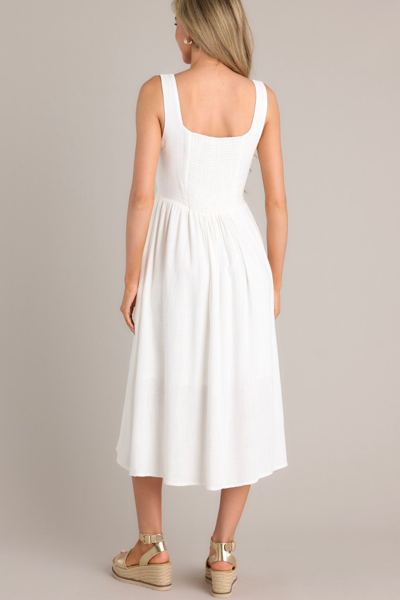 Back view of this white dress with a square neckline, thick straps, smocked back insert, side zipper, waist gathering, and hip pockets.