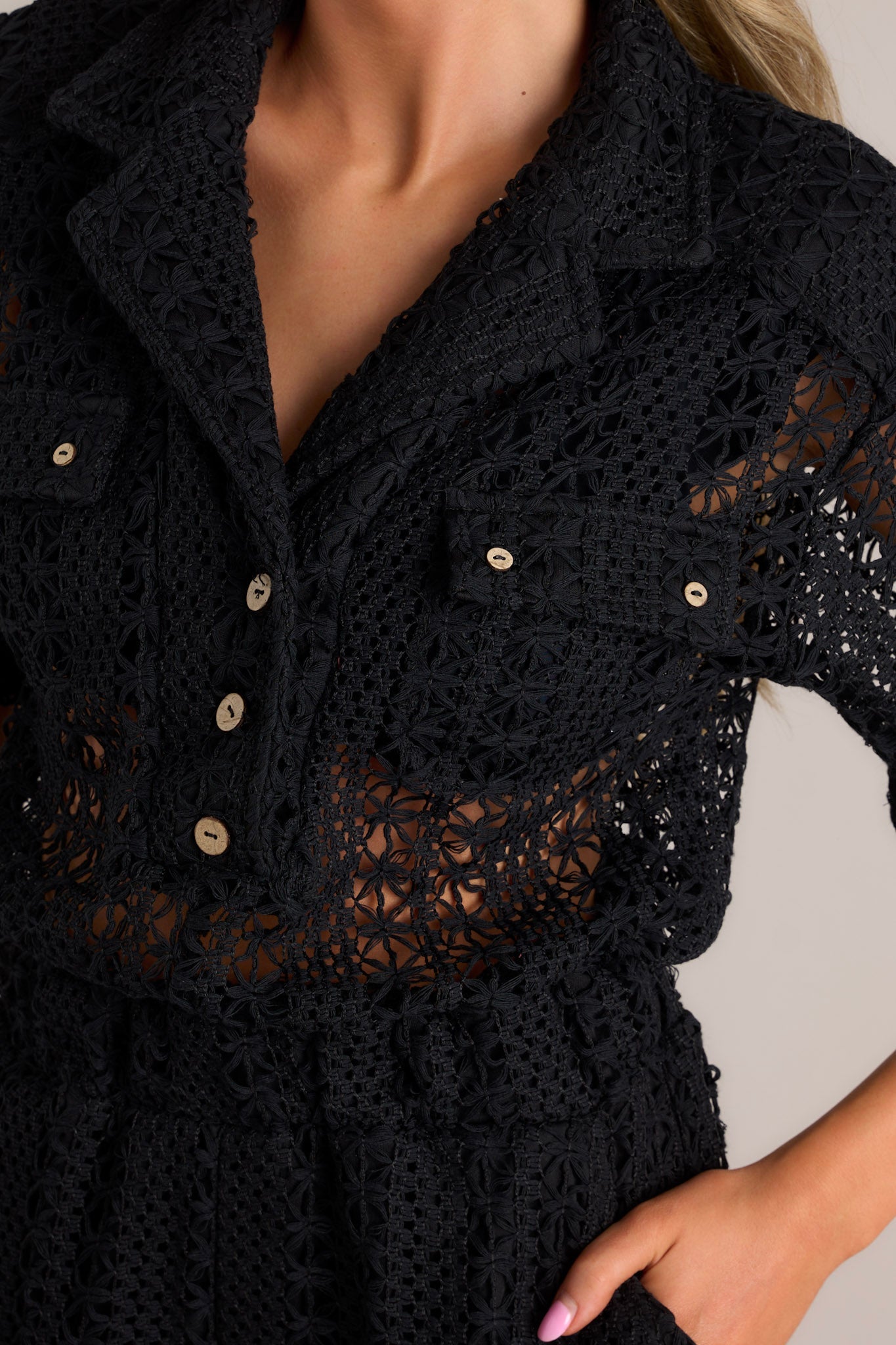 Close up view of this black open knit romper that features a collared neckline, functional buttons, faux breast pockets, an open knit design, a fitted waistband, and functional hip pockets.