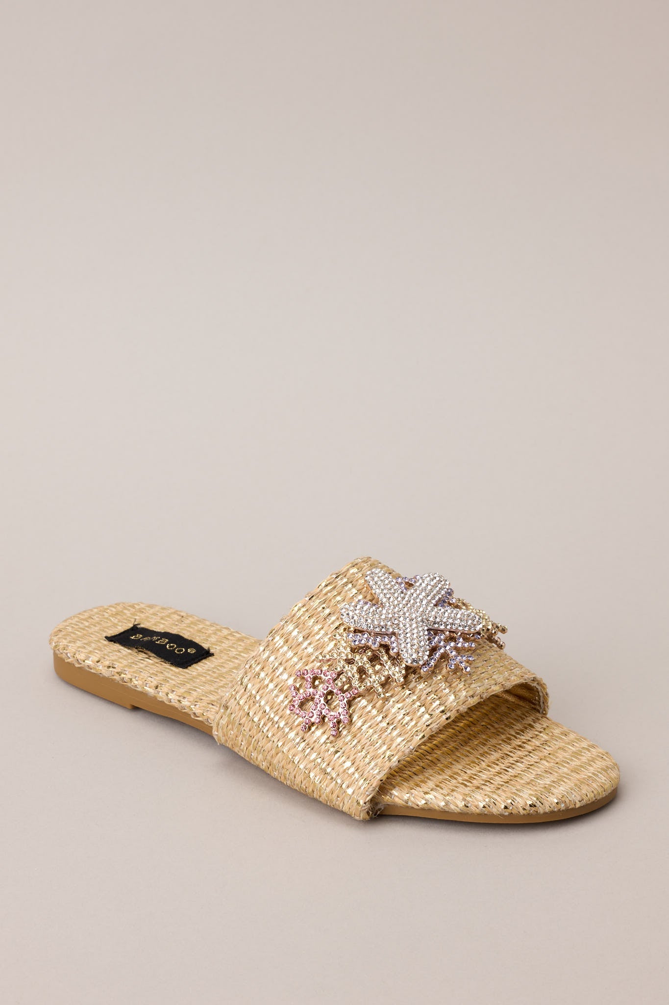 Angled front view of these sandals that feature a rounded toe, a slip-on design, a wicker-like material, and tropical multicolored rhinestone detailing.