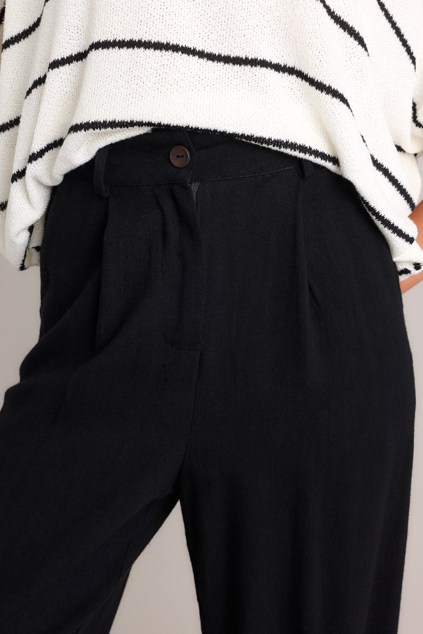 Close up view of these black linen joggers that feature a high rise design, a functional button & zipper closure, belt loops, subtle pleats, functional front & back pockets, and elastic cuffed ankles