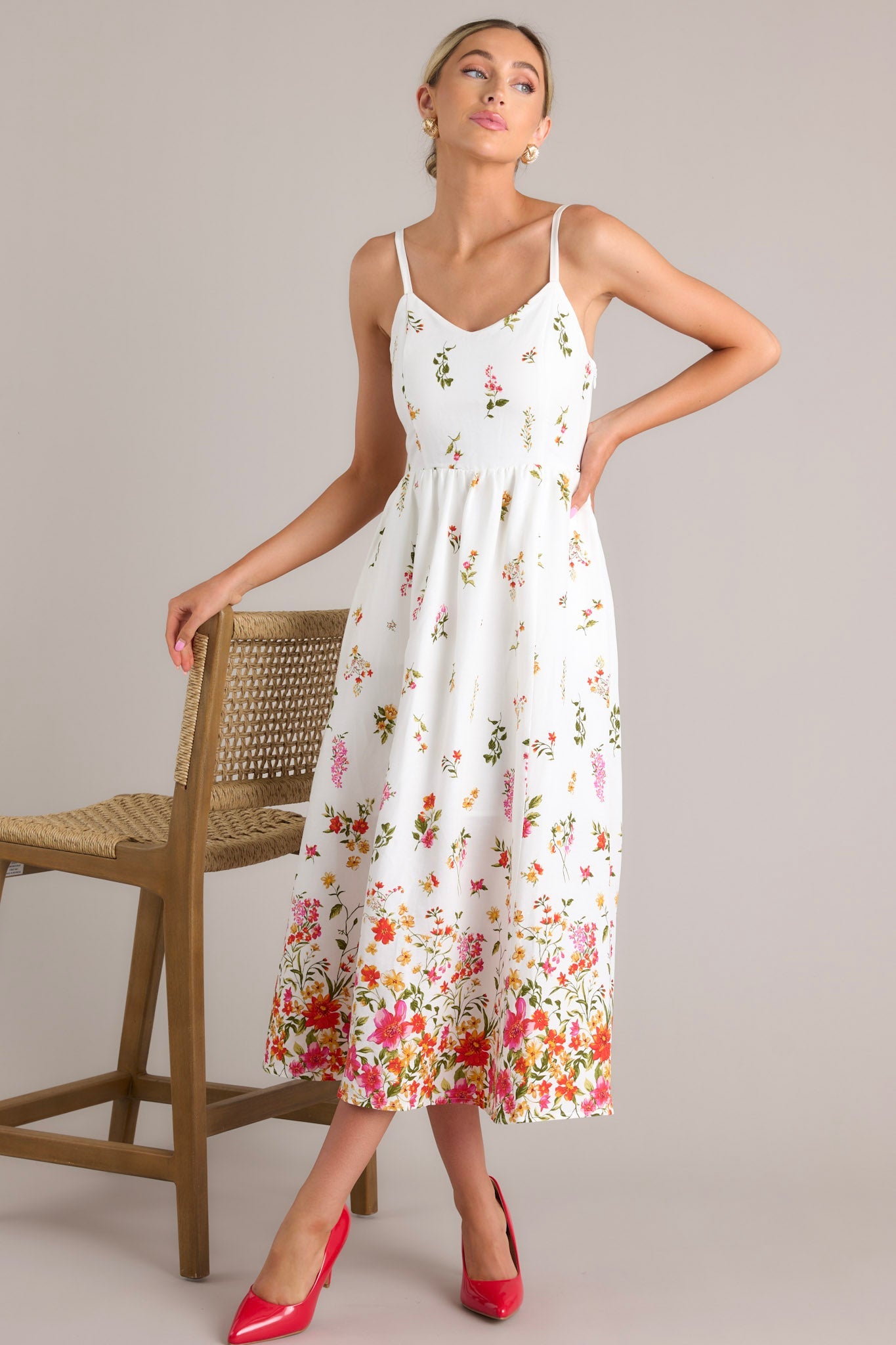 Ivory floral midi dress with thin adjustable straps and a fitted bodice, featuring a vibrant botanical print near the hemline, paired with red heels.