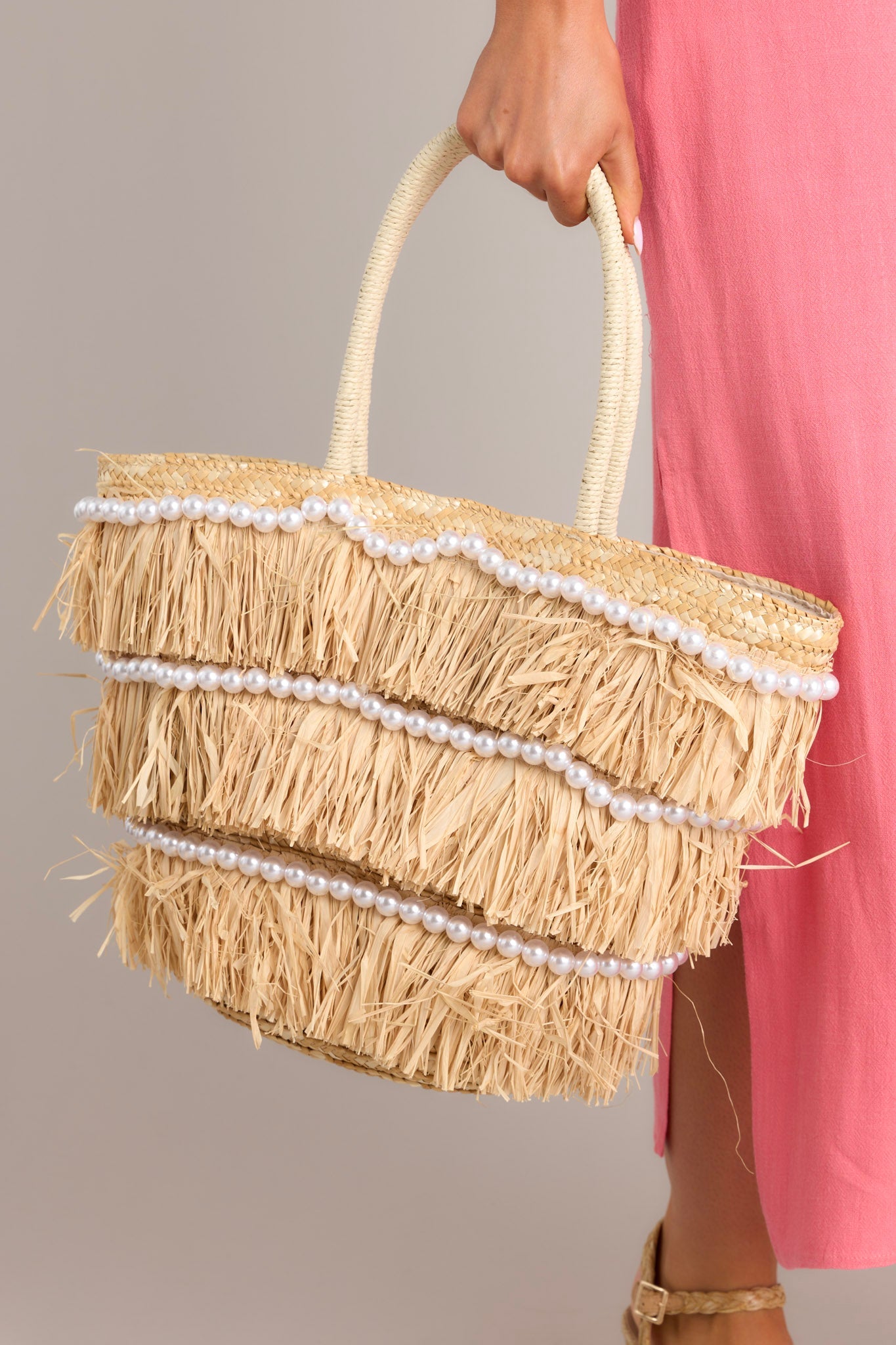 Close up view of this tote bag that features a woven designed, straw fringe detailing, rows of pearls, and double woven handles