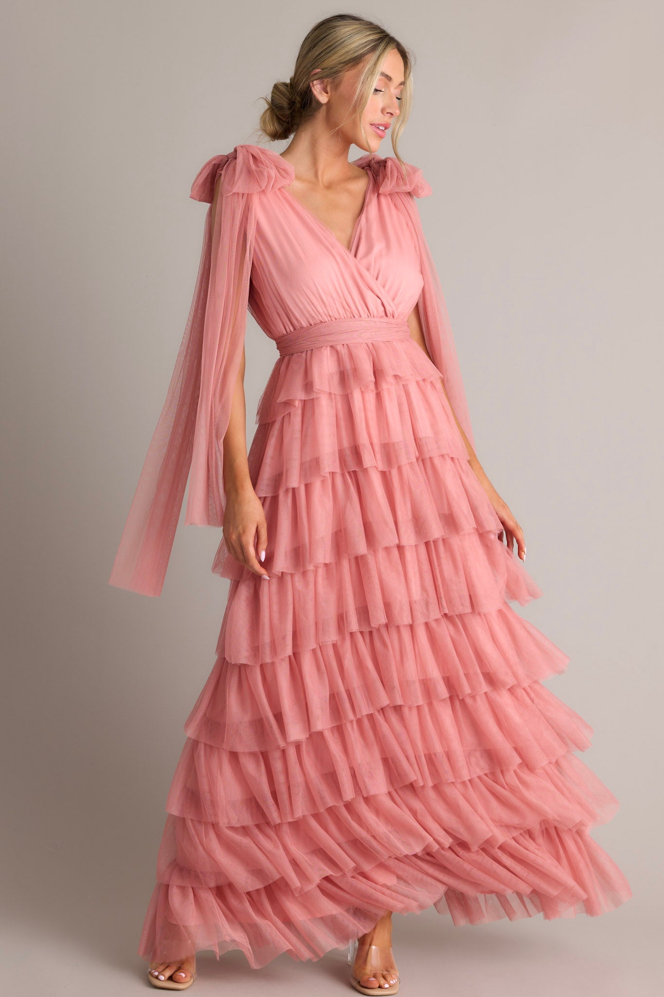 Pink dress featuring a flattering v-neckline, graceful fabric trailing from the shoulders, a chic self-tie waist belt, and multiple tiers and layers of ethereal tulle for a whimsical touch.