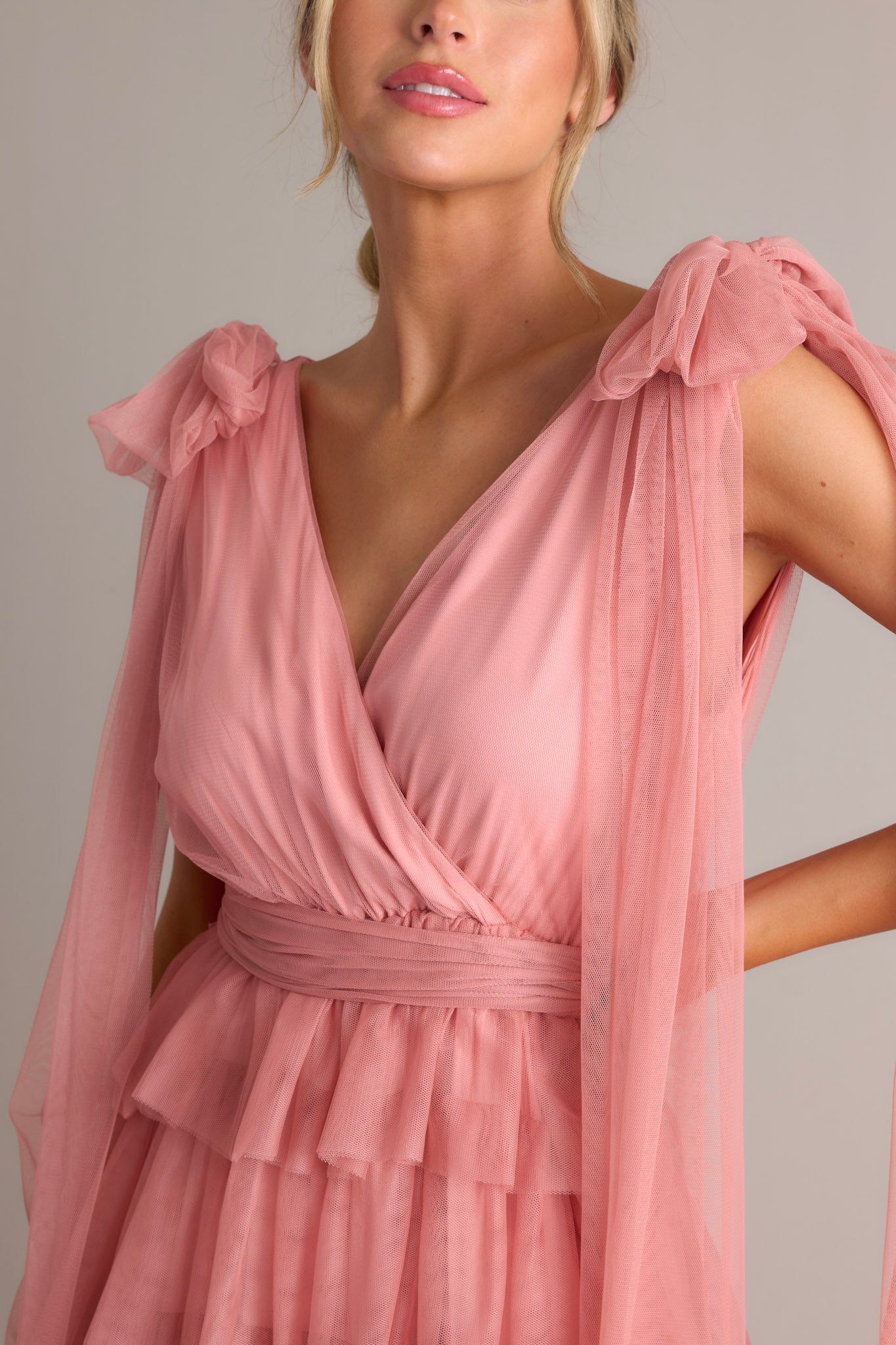 Close up view of this pink dress featuring a flattering v-neckline, graceful fabric trailing from the shoulders, a chic self-tie waist belt, and multiple tiers and layers of ethereal tulle for a whimsical touch.