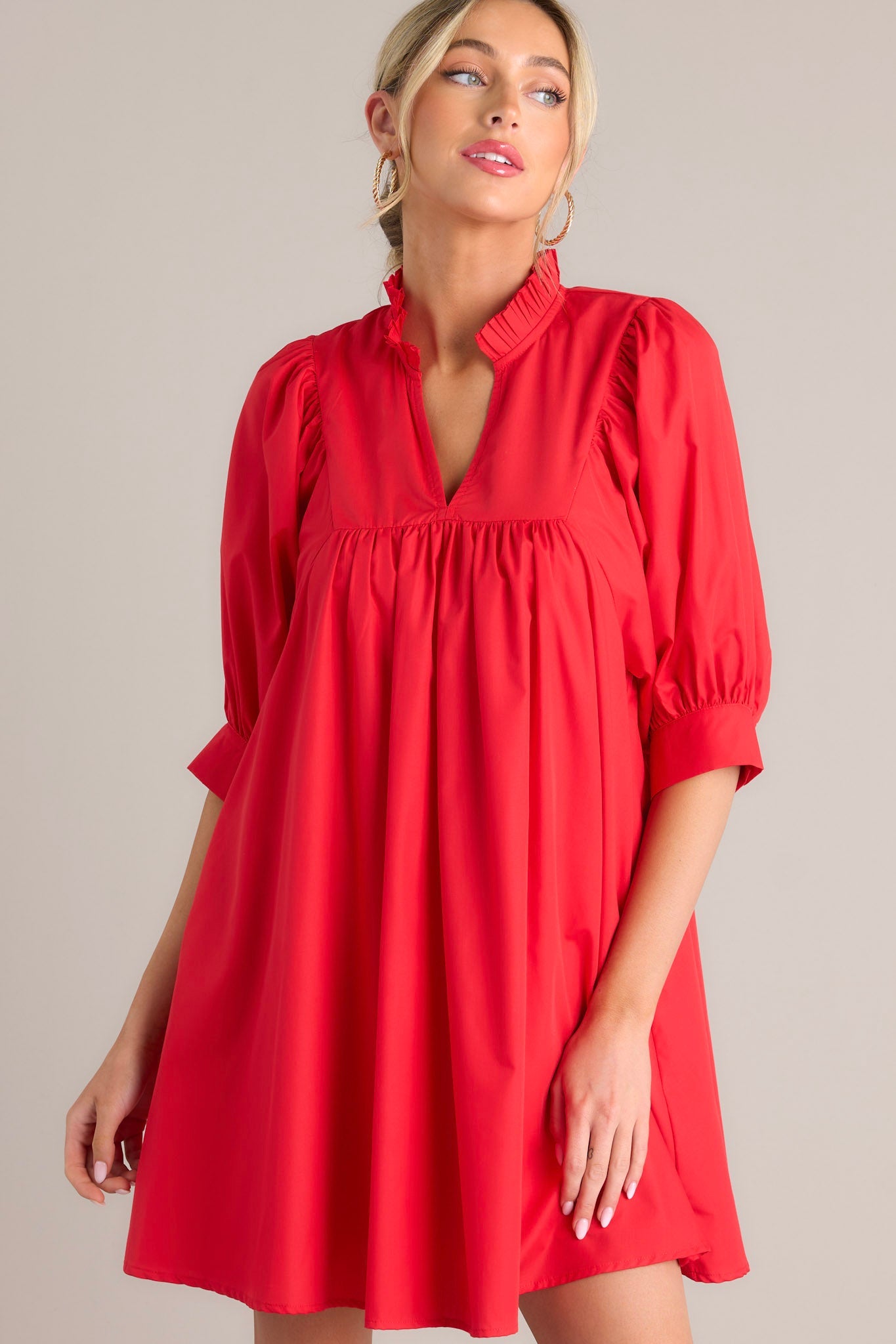 Angled front view of this red mini dress that features a v-neckline, a ruffled faux collar, pleated detailing around the bust, functional hip pockets, and cuffed puff sleeves.