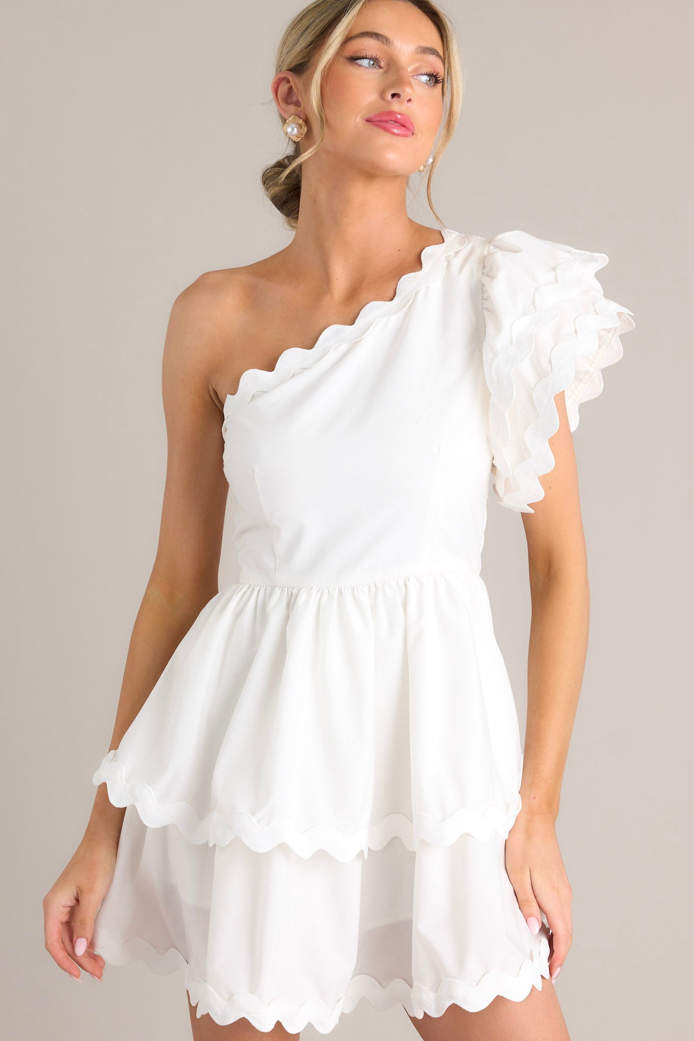 Front angled view of a white romper featuring rick rack scalloped hems, a one shoulder neckline, a flutter sleeve, a side zipper closure, and a tiered skirt with built-in shorts