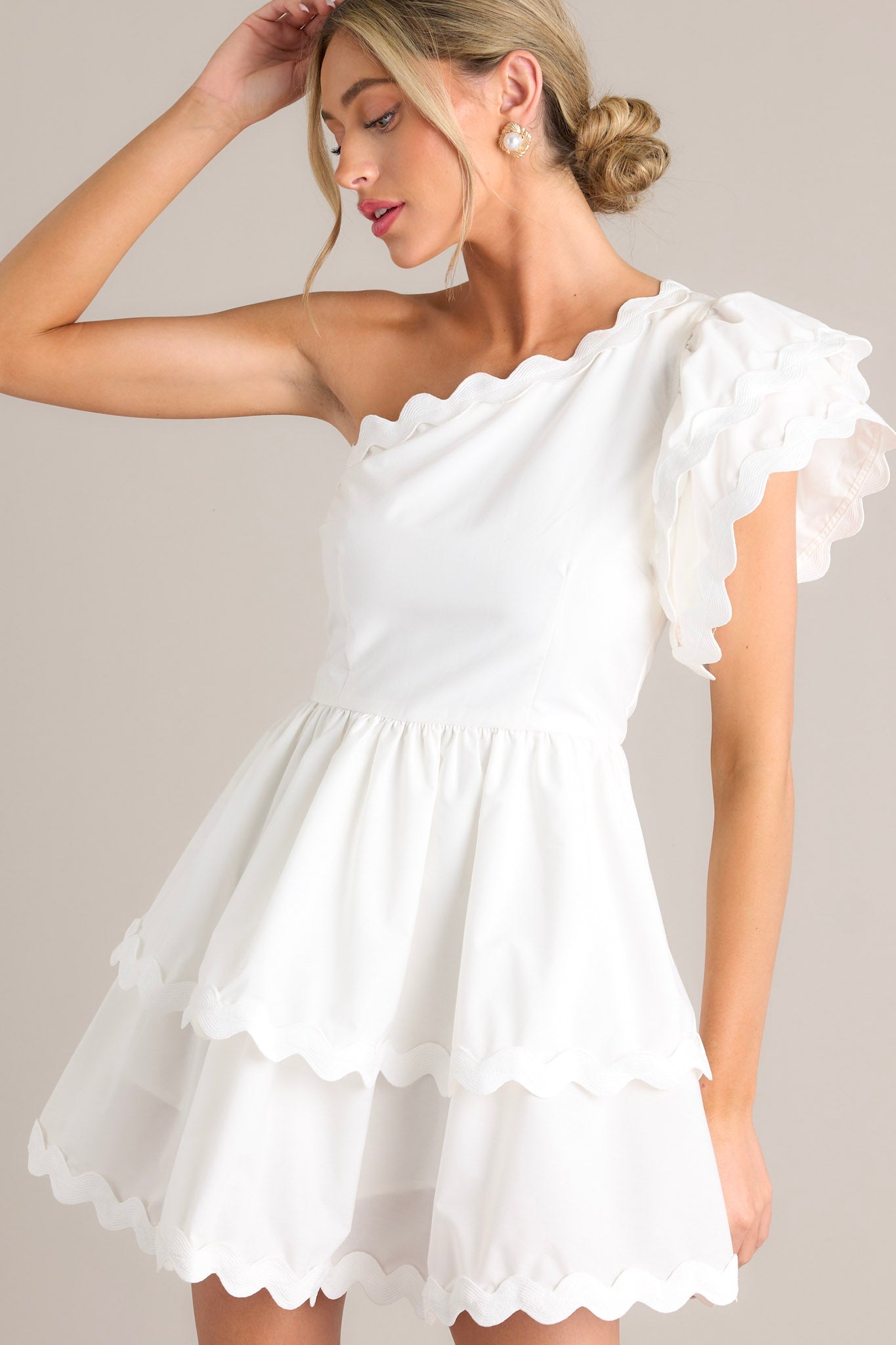Front view of a white romper featuring rick rack scalloped hems, a one shoulder neckline, a flutter sleeve, a side zipper closure, and a tiered skirt with built-in shorts.
