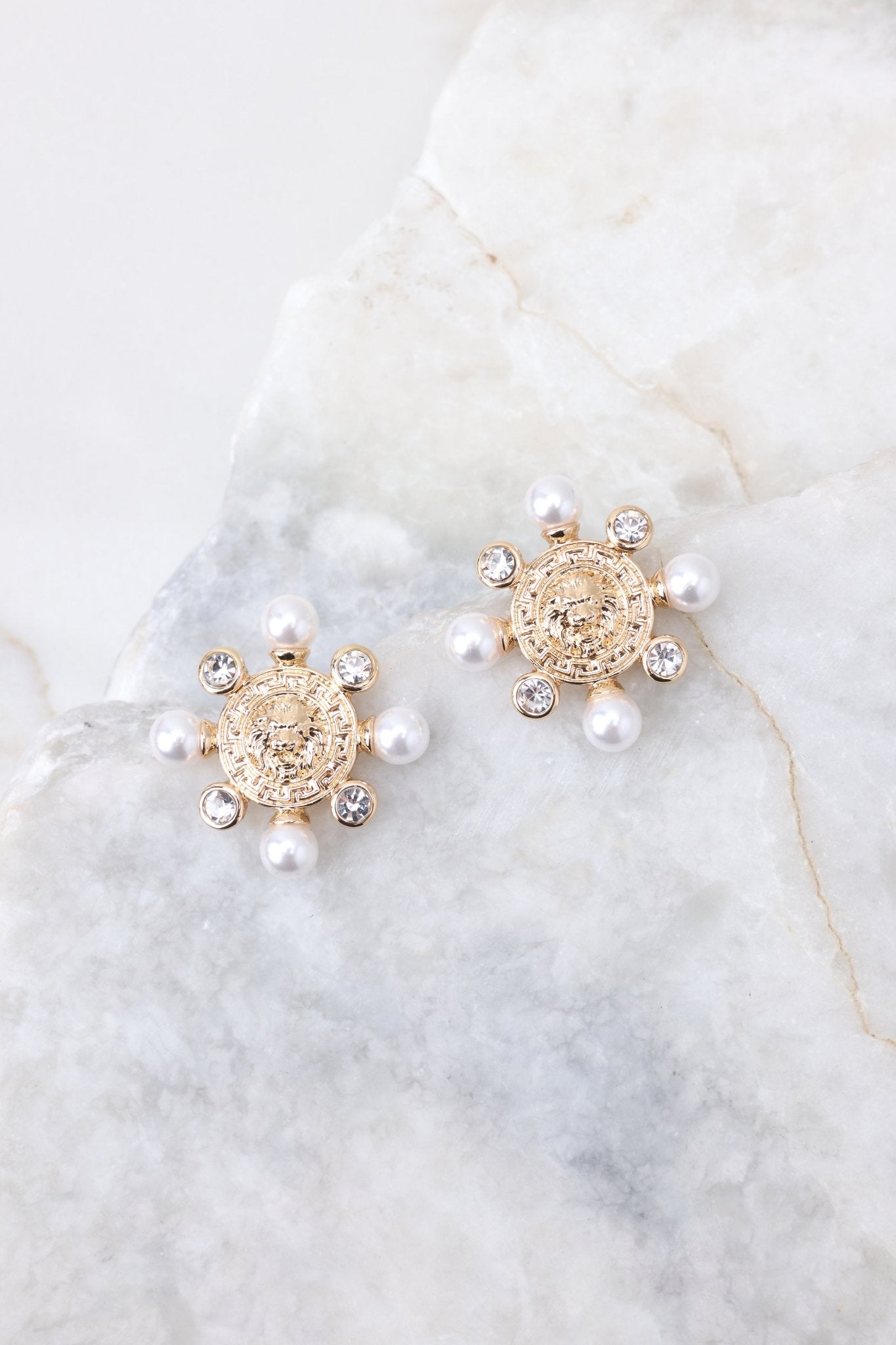 Overhead view of these earrings that feature a circle lion head design, small pearls and rhinestones along the edge, and a secure post backing.