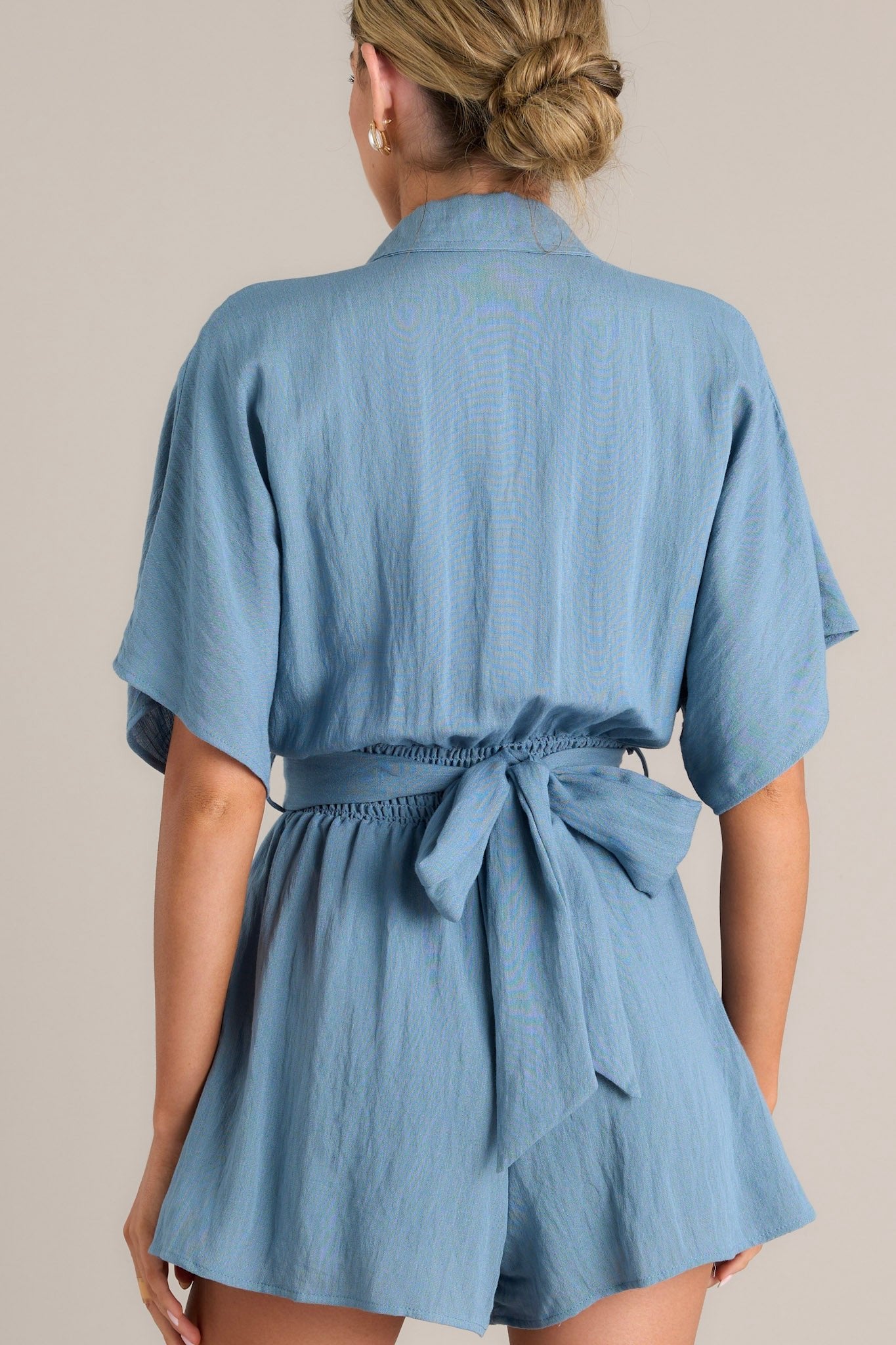 Back view of this ash blue romper that features a collared neckline, a button front chest, belt loops, a self-tie waist feature, and a smocked insert at the back of the waist.