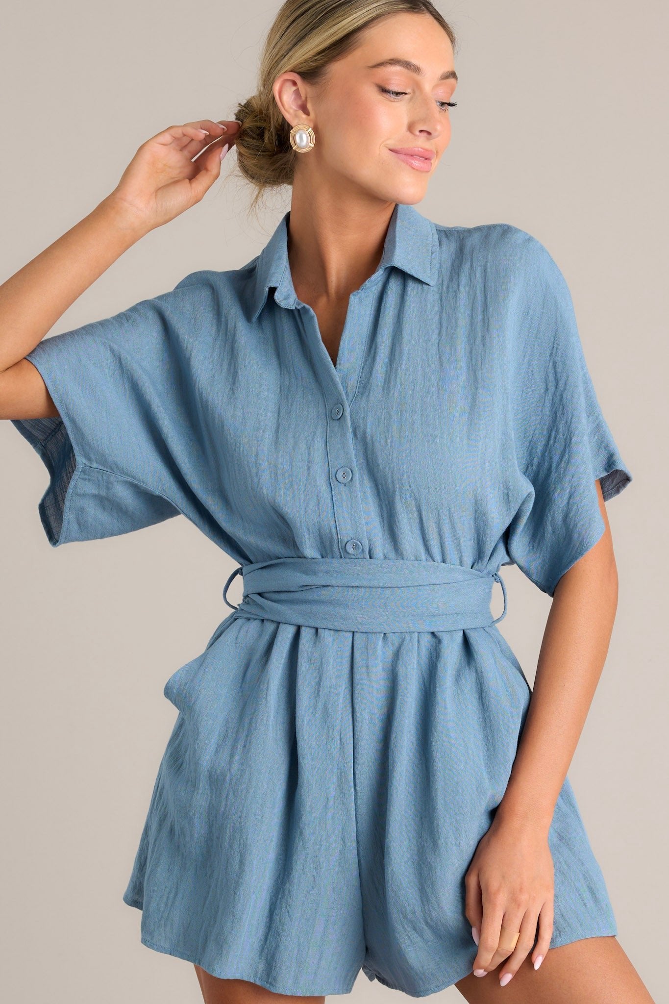 This ash blue romper features a collared neckline, a button front chest, belt loops, a self-tie waist feature, and a smocked insert at the back of the waist.