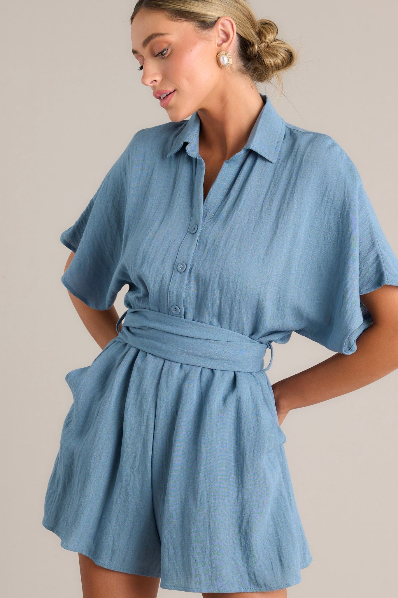 Angled close up view of this ash blue romper that features a collared neckline, a button front chest, belt loops, a self-tie waist feature, and a smocked insert at the back of the waist.