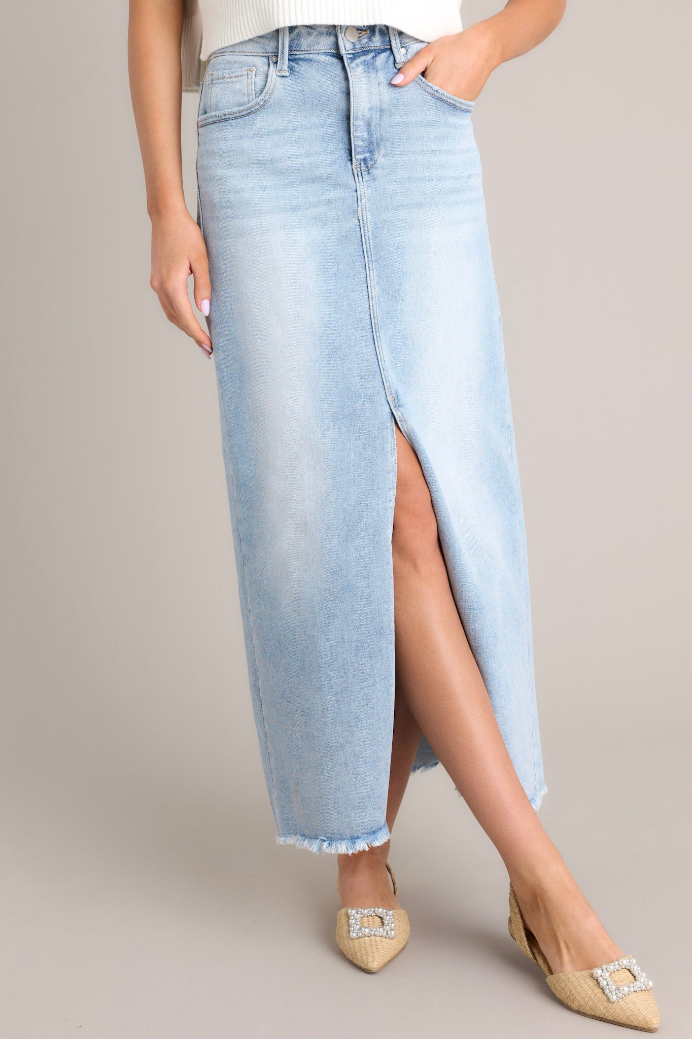Front angled view of a light wash denim midi skirt featuring a high waisted design, belt loops, a zipper and button closure, functional pockets, a 15" slit up the center, and raw hem detailing