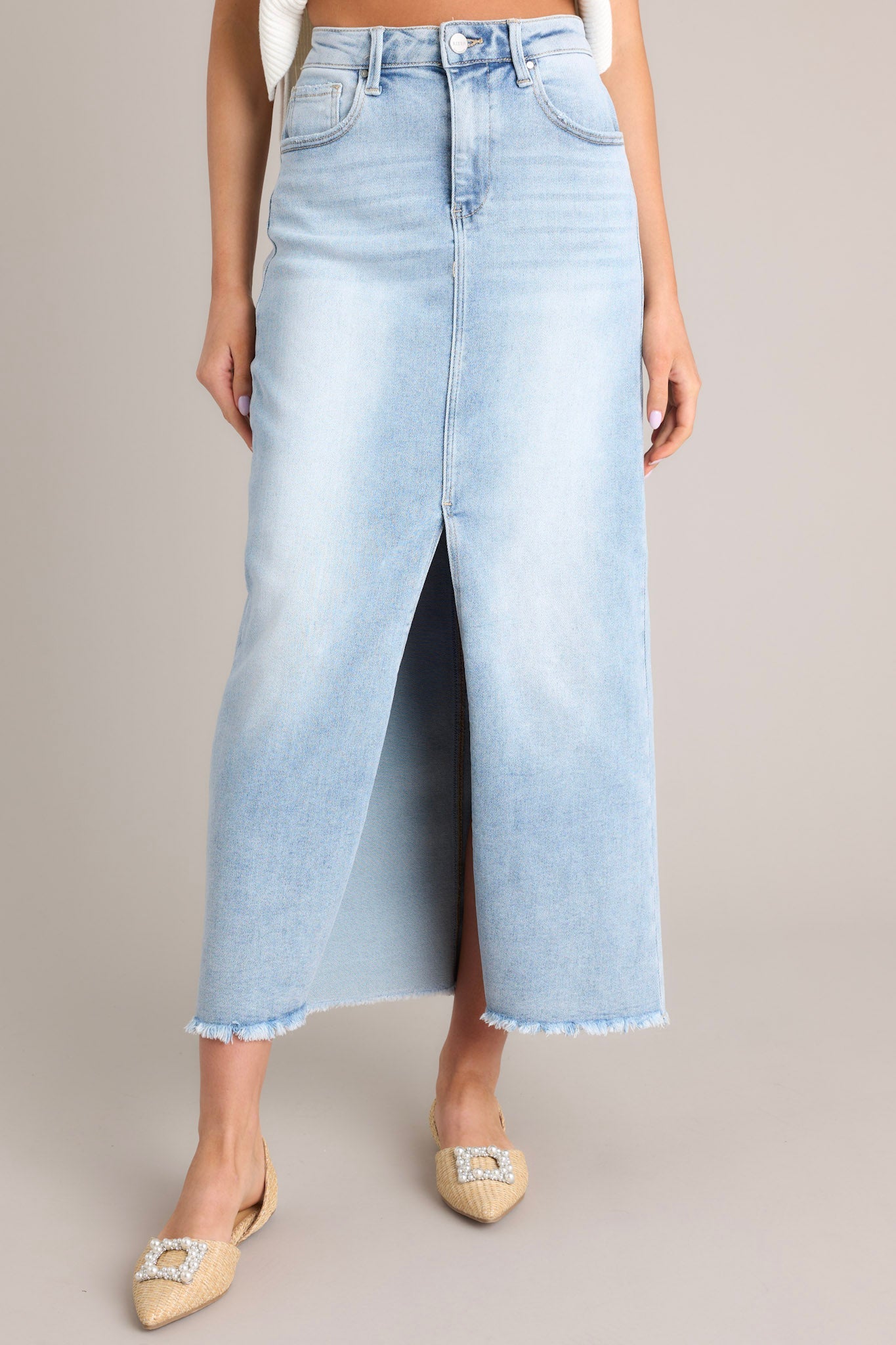 Front view of a light wash denim midi skirt featuring a high waisted design, belt loops, a zipper and button closure, functional pockets, a 15" slit up the center, and raw hem detailing.