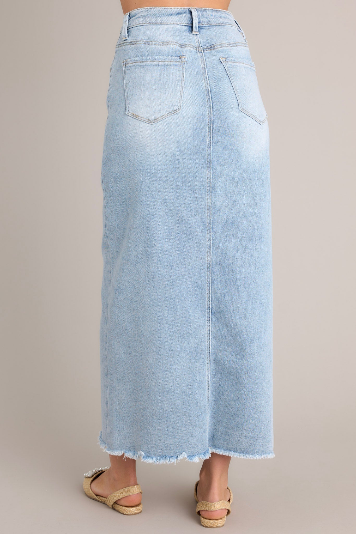 Back view of a light wash denim midi skirt highlighting the belt loops, functional back pockets, and raw hem detailing.