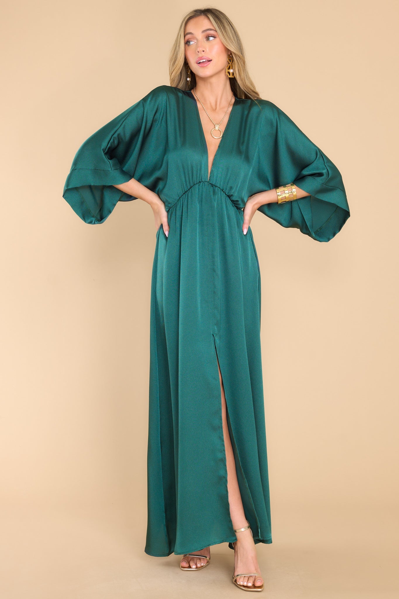 Full body view of this dress that features a plunging v-neckline, dolman sleeves, an elastic waistband, a flowy skirt with a slit up the center, and a lining that extends from the waist to the thigh.