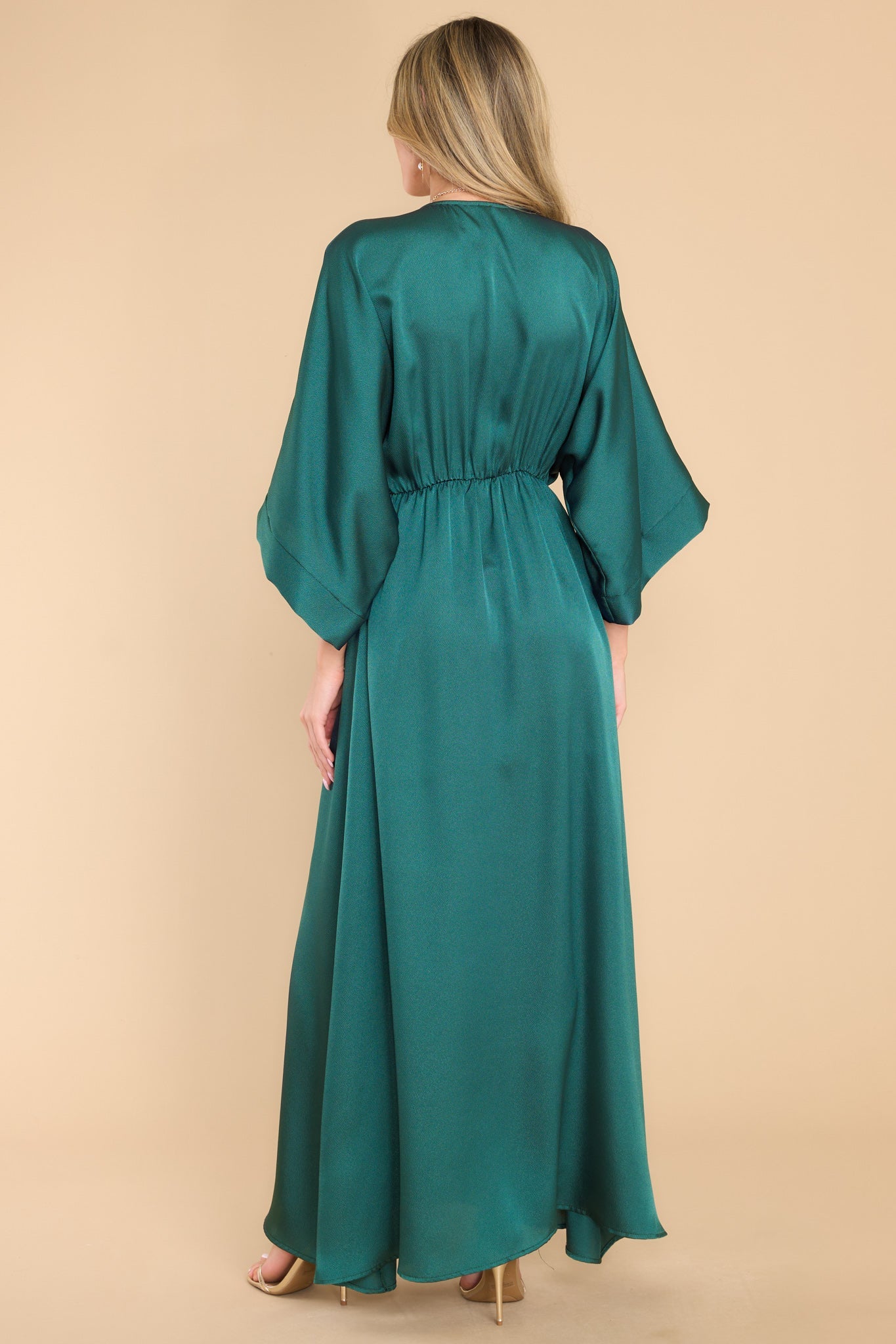 Back view of this dress that features a plunging v-neckline, dolman sleeves, an elastic waistband, a flowy skirt with a slit up the center, and a lining that extends from the waist to the thigh.