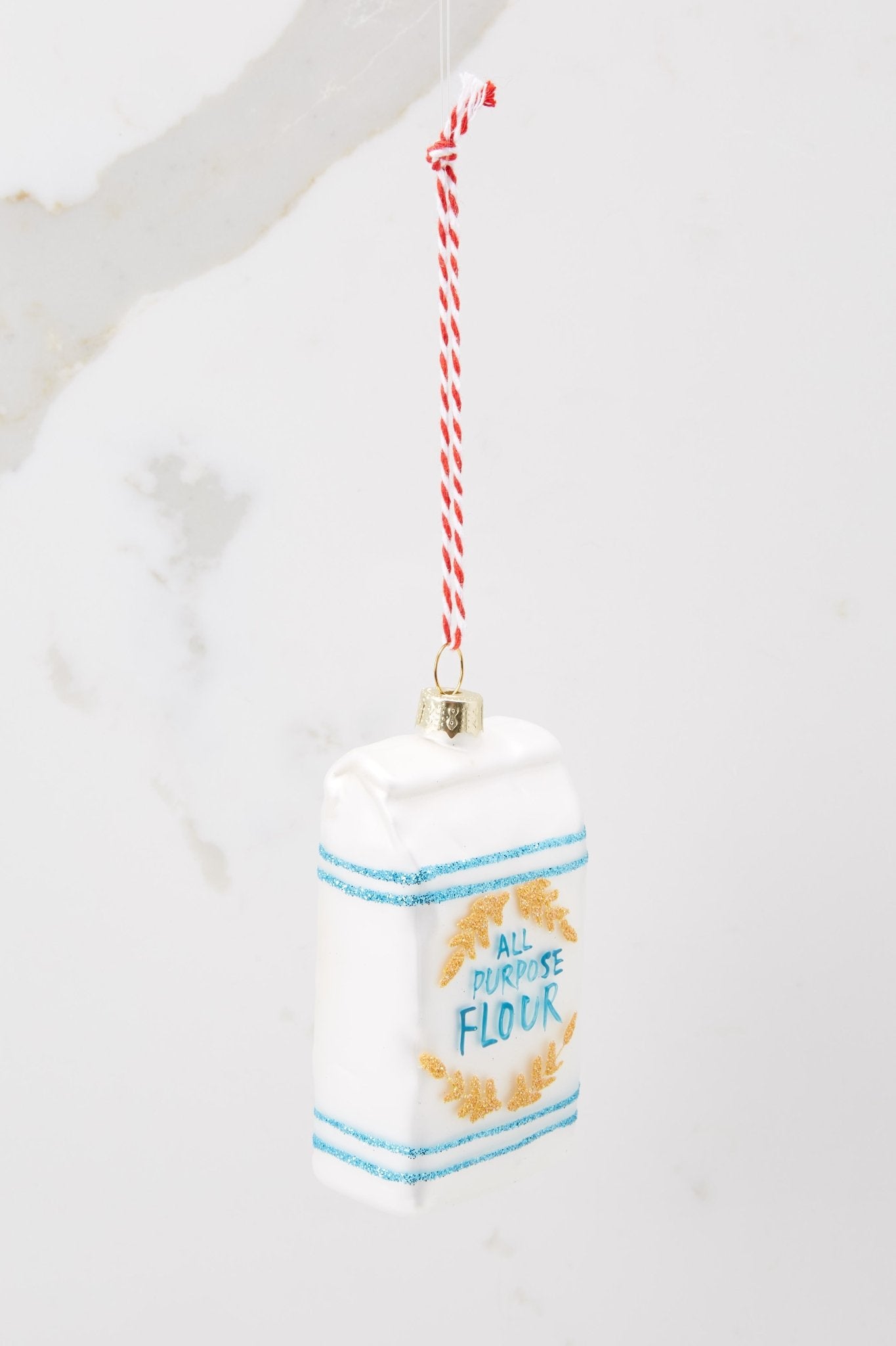 Angled side view of this ornament that features a design of 'all purpose' flour with hand painted blue lettering and gold glitter accents.