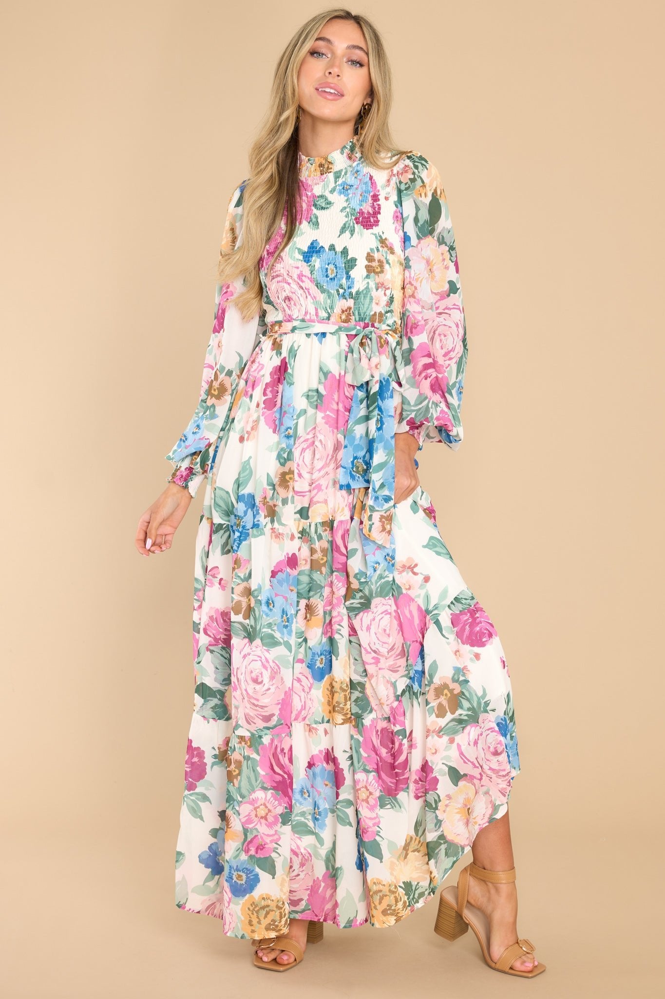 This ivory floral dress features a high neckline, a keyhole cutout at the back of the neck with a button closure a smocked bodice, sheer long sleeves with smocked cuffs, a self-tie belt at the waist, and a flowy skirt.