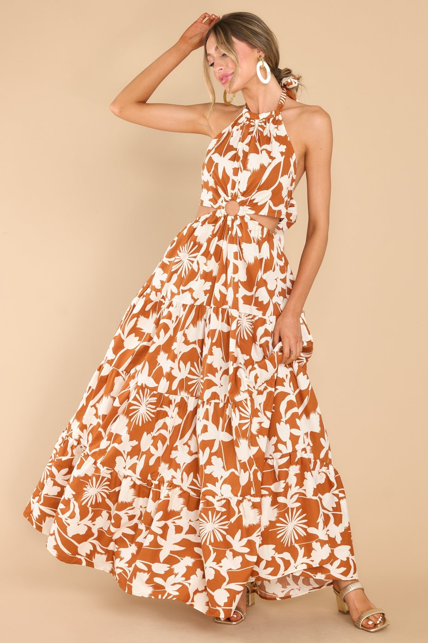 This brown and ivory dress features a halter neckline with adjustable self tie around the neck and back, cut out detailing on the waist with a ring detail in the middle, elastic band around the waist, and a long flowy tiered skirt.